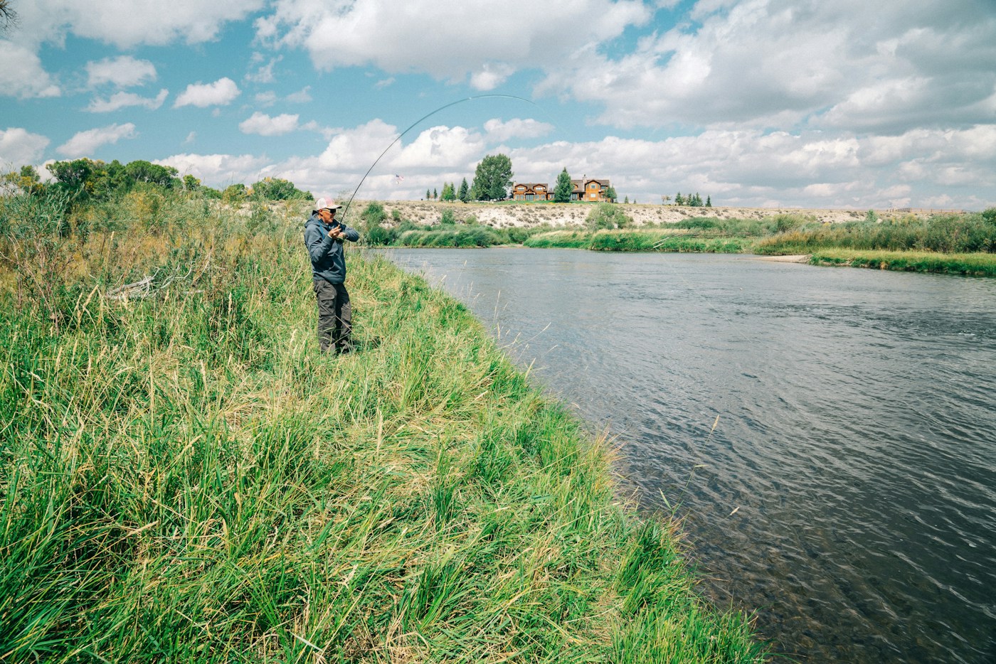 An angler casting a fishing rod from a grassy riverbank